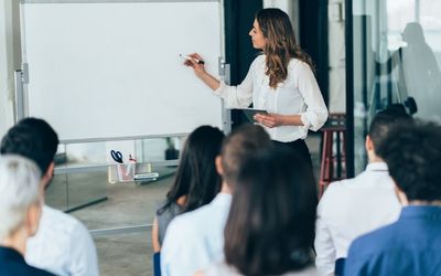 A female coach stands at a whiteboard leading an interactive group workshop session.